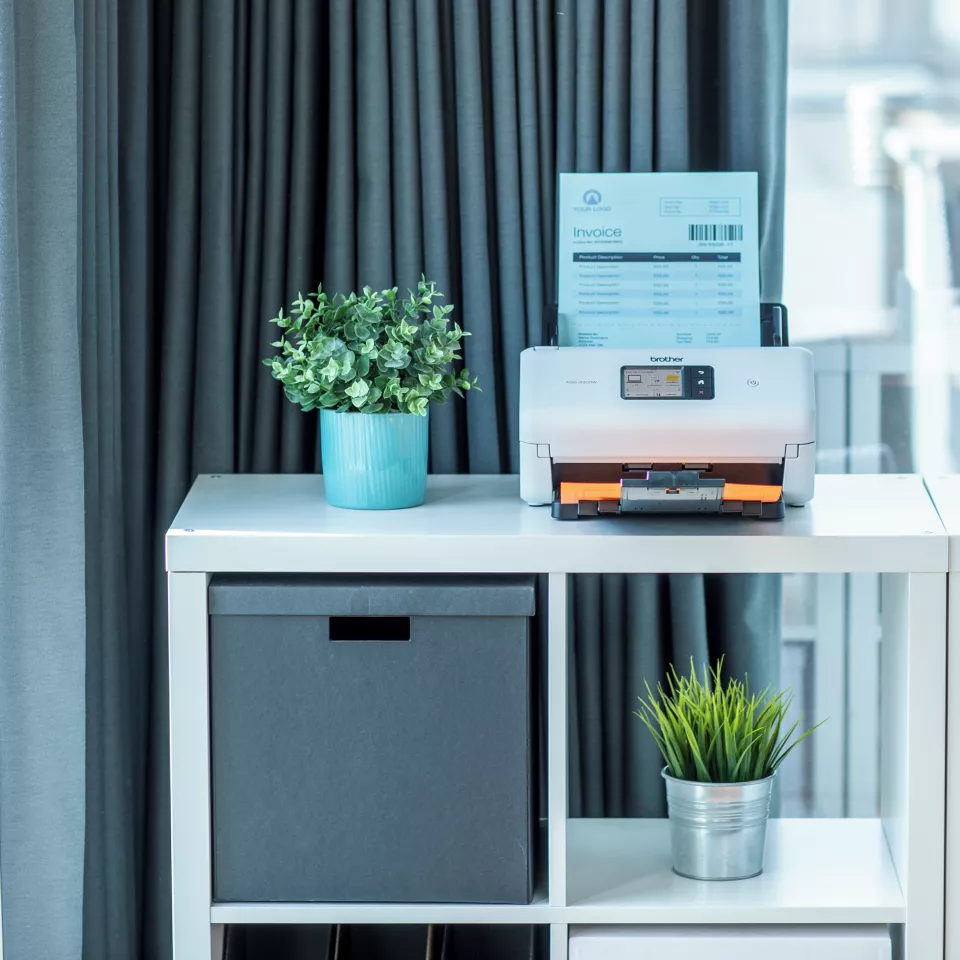 ADS-3300W sitting on a desk next to a pot plant with paper in the feeder and the tray